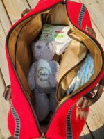 An open red and tan bag with brown handles containing baby items, including a package of Huggies Little Snugglers diapers, a soft gray teddy bear with "Destiny" embroidered on it, and additional folded diapers. The bag is placed on a wooden surface.