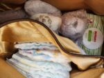 A brown bag contains several diapers, a pack of Huggies baby wipes, and two stuffed animals—one gray and one cream-colored. The contents appear neatly arranged, ready for use.