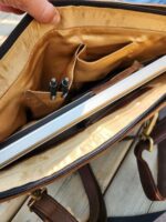 A close-up view of the interior of an open brown leather bag, revealing a compartment holding a laptop, two pens in slots, and a pocket in a gold-colored lining. A hand is grasping the upper part of the bag's opening.