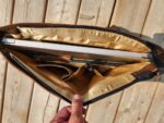A hand holds open a brown leather bag on a wooden surface. Inside the bag are a silver laptop, a pen, and various compartments made of gold fabric lining. Sunlight casts shadows on the items and the wooden background.