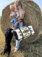 A woman stands in front of a hay bale holding a baby on her hip and a patterned bag in her other hand. A small black dog stands on its hind legs, paws resting on the woman's leg, looking up at the baby. They are outdoors on a sunny day.