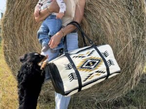 A woman stands in front of a hay bale holding a baby on her hip and a patterned bag in her other hand. A small black dog stands on its hind legs, paws resting on the woman's leg, looking up at the baby. They are outdoors on a sunny day.