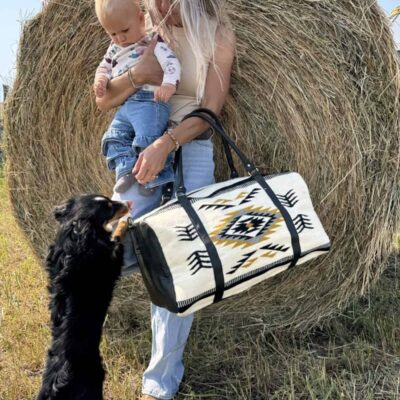 A woman stands in front of a hay bale holding a baby on her hip and a patterned bag in her other hand. A small black dog stands on its hind legs, paws resting on the woman's leg, looking up at the baby. They are outdoors on a sunny day.