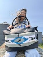 A person with long hair, partially obscured under a hood, holds a large tote bag with a Native American-inspired geometric pattern in blue, white, and black. They're seated on a straw bale with silos visible in the background under a blue sky.