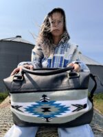 A person with long hair, partially obscuring their face, sits outdoors on a bale of hay. They wear a patterned hoodie and hold a large black bag featuring a tribal design with blue, white, and black colors. Metal silos are visible in the background under a clear sky.