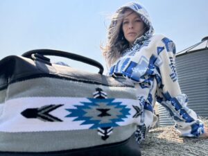 A person wearing a hooded garment with a blue and white geometric pattern sits atop a hay bale under a clear sky. In the foreground, there is a bag with a similar geometric pattern in blue, white, and black. Their expression is neutral.