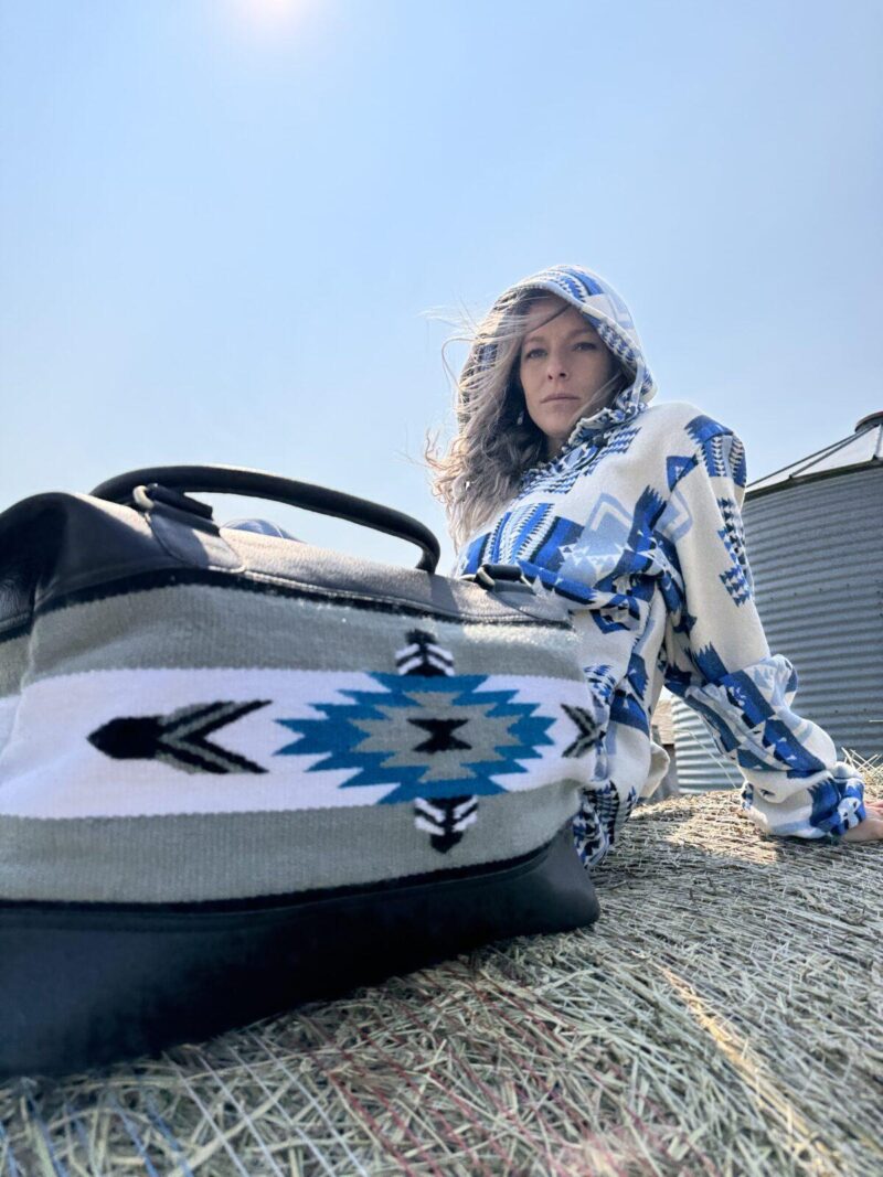 A person wearing a hooded garment with a blue and white geometric pattern sits atop a hay bale under a clear sky. In the foreground, there is a bag with a similar geometric pattern in blue, white, and black. Their expression is neutral.