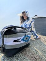 A person wearing a blue-and-white patterned hoodie sits outdoors on a hay bale. A large bag with a similar geometric pattern is placed in the foreground. The background shows a clear blue sky and part of a metal structure. The person's hair is being blown by the wind.