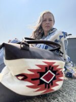 A person with long hair is sitting outdoors on a hay bale, wearing a blue and white patterned jacket. In the foreground, there is a large woven bag with a geometric red, black, and white design and leather accents. The sky is clear.