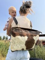 A person with light brown, messy hair is holding a baby with blonde hair. The person is wearing a beige top, light blue denim jeans, and a brown and white cowhide shoulder bag. They are standing in a grassy field with a clear blue sky in the background.