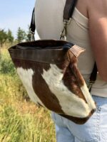 A person is wearing a brown and white cowhide bag over their shoulder, standing in a grassy field. The bag has a leather strap and metal clasps. The person is dressed in a white tank top and light blue pants. Trees are visible in the background.