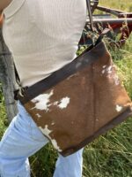 A person wearing a white sleeveless shirt and light blue jeans is standing outdoors beside a fence. They are carrying a brown and white cowhide bag with a leather strap over their shoulder. The background features grass and blurred farm equipment.