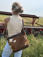 A person with light blonde hair in a messy bun stands in a grassy field, wearing a beige tank top, blue jeans, and a brown leather bag with white patches over their shoulder. They are facing away from the camera towards a red farming machine in the background.