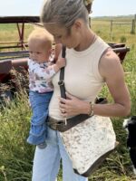 A woman with blonde hair in a ponytail, wearing a sleeveless beige top and light blue jeans, holds a baby dressed in a floral top and blue pants. They are outdoors in a grassy area, with part of a red farm vehicle visible in the background. A black and white dog is at their feet.