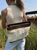 A person with long hair in a ponytail, wearing a beige tank top and light blue jeans, stands in a grassy field. They carry a brown and white spotted cowhide bag over their shoulder. In the background, there's agricultural machinery and hay bales.