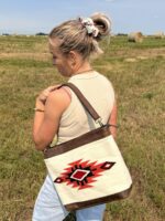 A woman with a bun hairstyle stands in a grassy field, looking away from the camera. She carries a shoulder bag with a brown strap and a Southwest-inspired geometric pattern in red, black, and white. Hay bales and a clear sky are visible in the background.