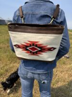 A person wearing a denim jacket and jeans is shown from the back, carrying a shoulder bag with a Native American-inspired geometric design. The bag has brown leather accents and a red, black, and white pattern. A small black dog is sniffing the grass nearby.