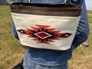 A person wearing a denim jacket and jeans is shown from the back, carrying a shoulder bag with a Native American-inspired geometric design. The bag has brown leather accents and a red, black, and white pattern. A small black dog is sniffing the grass nearby.