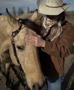 A person wearing a cowboy hat, brown jacket, and scarf is gently patting the head of a tan horse wearing a bridle. With long hair and a blue ring on their middle finger, they share a calm moment outdoors with trees and a hill in the background, feeling at home in nature’s embrace.