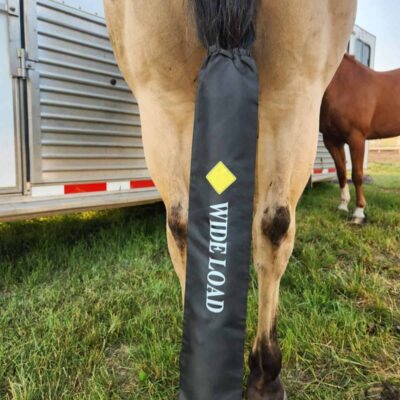 A close-up of a horse's hindquarters with a black cover over its tail. The cover has the words "WIDE LOAD" and a yellow diamond symbol on it, indicating a cautionary message. Another horse and a trailer can be seen in the background.