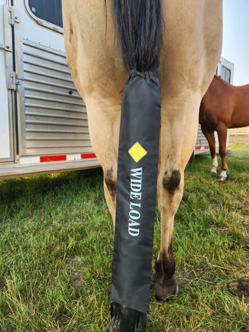 A close-up of a horse's hindquarters with a black cover over its tail. The cover has the words "WIDE LOAD" and a yellow diamond symbol on it, indicating a cautionary message. Another horse and a trailer can be seen in the background.