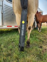 A horse standing in a grassy area with a "WIDE LOAD" sign cover attached to its tail. In the background, another horse and a person wearing a hat are visible, along with a horse trailer.