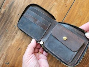 A person holding an open Wallet - Slim with brown leather tri-fold design and a brass button closure on a wooden table. The wallet, perfect as a gift, features multiple card slots on the left side and a coin pouch on the right side.