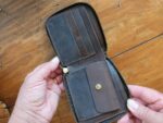 A person is holding open the Wallet - Slim with, crafted from brown leather in a tri-fold design featuring a zipper. The interior showcases multiple card slots on the left and a snap-button coin pocket on the right. This elegant accessory, ideal for gifting, is displayed against a wooden surface.