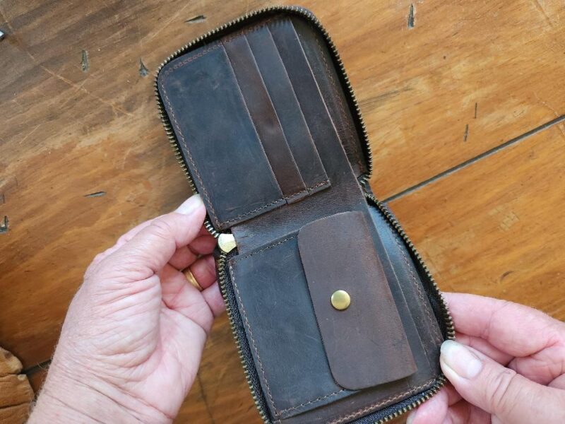 A person is holding open the Wallet - Slim with, crafted from brown leather in a tri-fold design featuring a zipper. The interior showcases multiple card slots on the left and a snap-button coin pocket on the right. This elegant accessory, ideal for gifting, is displayed against a wooden surface.