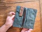 A person is holding an open army green canvas wallet with brown leather accents. The wallet, resembling a canvas clutch, features a snap button closure, a zippered pocket, and several card slots. The background is a wooden surface.