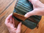 Close-up of a pair of hands holding and opening an army green canvas clutch with brown leather accents and a zipper. The item appears to have multiple compartments and is resting on a wooden surface.