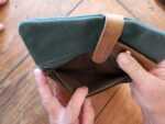 A person holds open a small, empty, army green canvas pouch with brown leather accents against a wooden surface background. The pouch's zipper is partially open, revealing its empty interior. Two fingers are visible, holding the brown leather strap of what could be used as a canvas clutch.