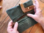 Two hands are holding an open army green canvas clutch with a brown leather strap and a zippered pocket. The wallet is placed on a wooden surface, while a third hand is partially visible in the top left corner of the image.