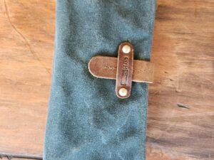 An army green canvas clutch with a brown leather strap and brass button closure lies on a wooden surface. The strap has the word "Standard" embossed on it.