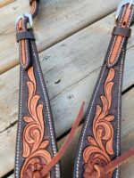 Close-up of two intricately designed leather belts with embossed floral patterns in shades of brown. Each belt features silver buckles and stitching along the edges. The belts are laid out on a wooden surface.