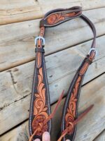 A dark leather belt with intricate floral designs and silver buckles is laid out on a wooden surface. The belt has light brown decorative stitching and leather straps hanging from it. One end of the belt is being held by a person's hand.