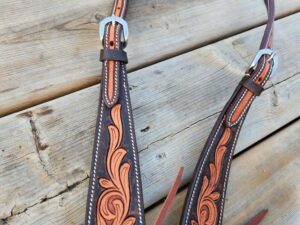 A dark leather belt with intricate floral designs and silver buckles is laid out on a wooden surface. The belt has light brown decorative stitching and leather straps hanging from it. One end of the belt is being held by a person's hand.