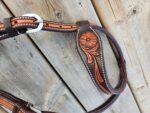 A close-up photo of a leather horse bridle with intricate floral tooling on a wooden surface. The leather is dark brown with a lighter brown floral pattern, white stitching, and a silver buckle.