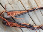 A close-up image of a leather horse bridle with intricate floral carvings. The bridle features a combination of dark and tan leather, with detailed stitching and laces. The background consists of wooden planks that have a weathered appearance.