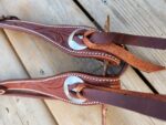 Close-up of two brown leather horse bridles with intricate stitching and silver embellishments, placed on a wooden surface. The bridles are detailed with carved patterns, showcasing careful craftsmanship.