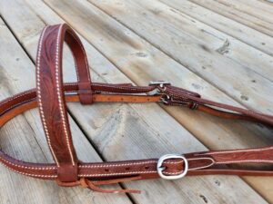 A close-up of a brown leather horse bridle with white stitching, resting on wooden planks. The bridle includes a buckle and several straps, displaying detailed craftsmanship. The wooden background has a rustic appearance.