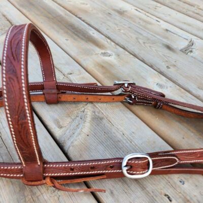 A close-up of a brown leather horse bridle with white stitching, resting on wooden planks. The bridle includes a buckle and several straps, displaying detailed craftsmanship. The wooden background has a rustic appearance.