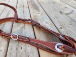 Close-up of a brown leather horse bridle with detailed white stitching and metal buckles, lying on a wooden deck. The leather shows intricate craftsmanship in its design.