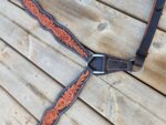 A close-up of a leather horse breast collar with intricate orange floral tooling and white stitching, resting on a wooden surface. The collar has metal hardware and adjustable straps.