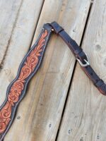 A detailed leather guitar strap lies on a wooden surface. The strap features intricate floral patterns in brown, bordered with light-colored stitching. It has a buckle adjustment for length customization.