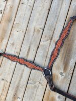 A leather harness with intricate, ornamental brown and black stitching is laid out on a wooden surface. The design features floral patterns and detailed craftsmanship, with adjustable straps visible on either side.